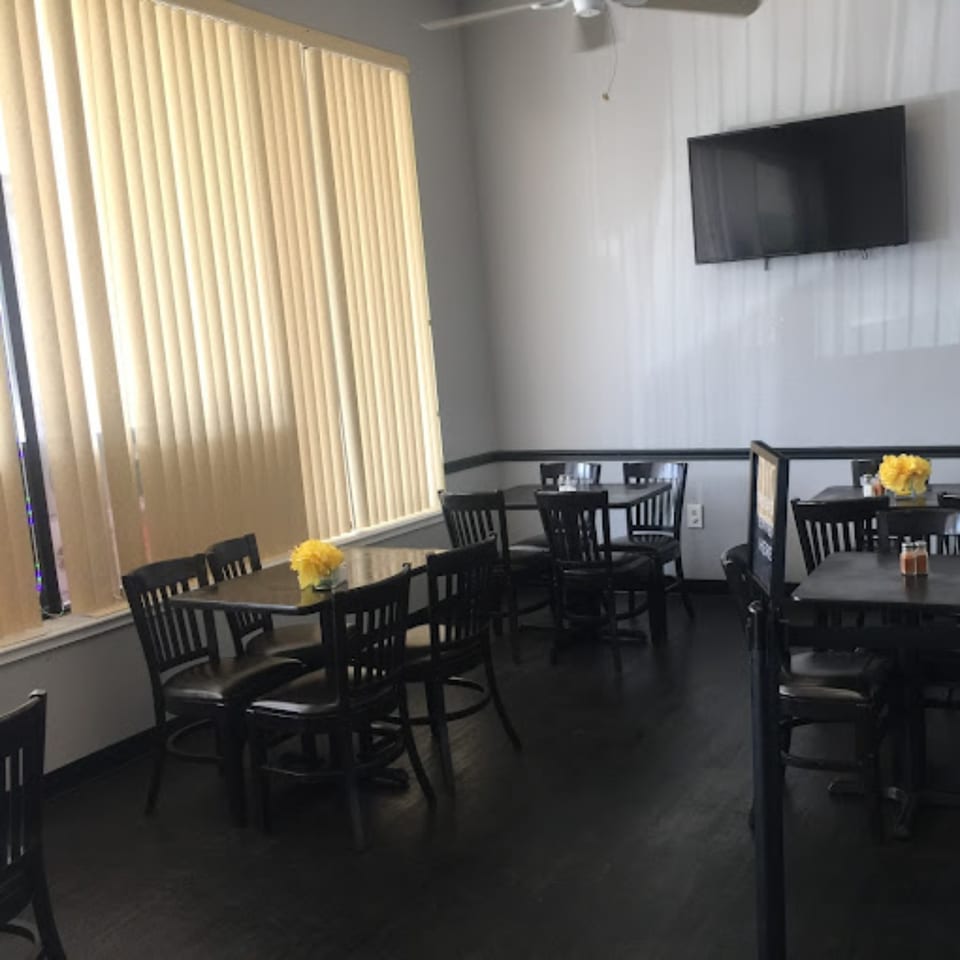 Interior of The Hunger Tamer restaurant with dark tables and chairs, light walls, and window blinds.