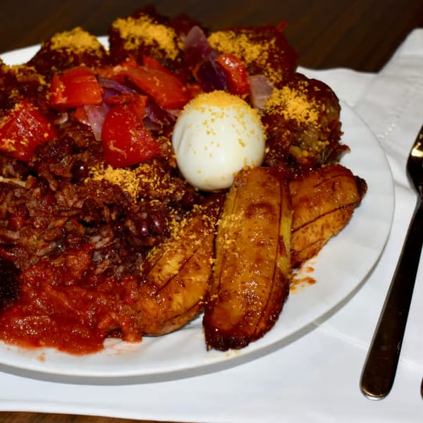 A plate of African cuisine with rice, plantains, stew, vegetables, and a hard-boiled egg.