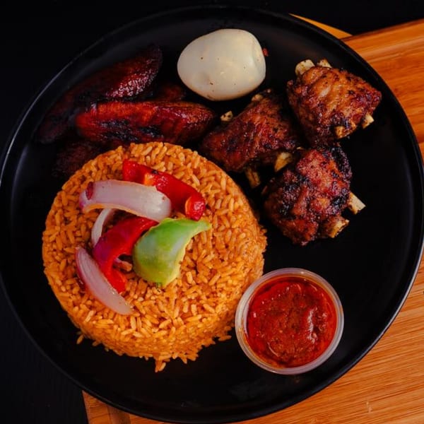 African jollof rice with grilled ribs, plantains, a boiled egg, and red sauce on a black plate.