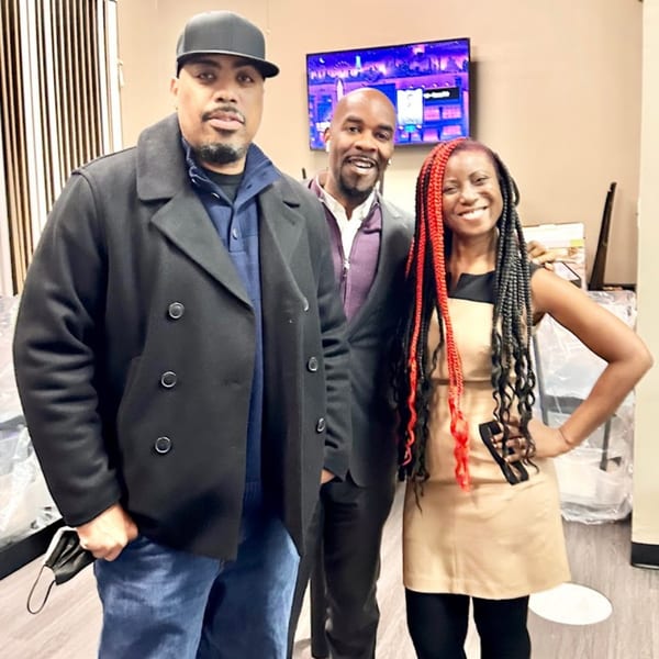 Three smiling people, two men and one woman with red and black braids, pose indoors.