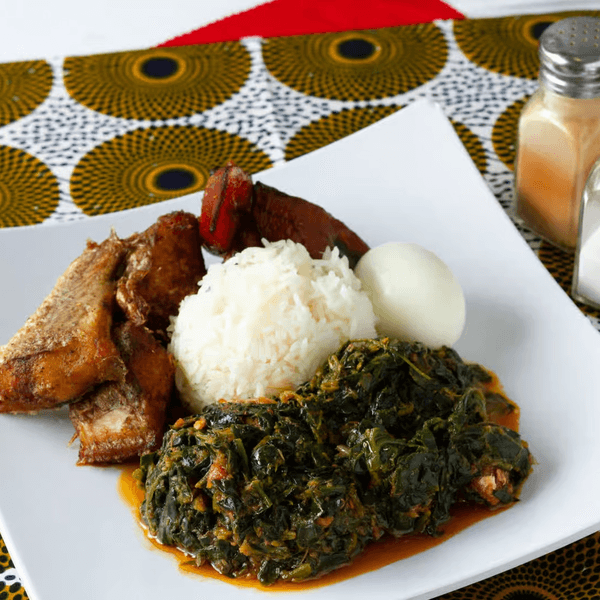African dish with fried fish, rice, stewed greens, plantain, and a hard-boiled egg on a white plate.